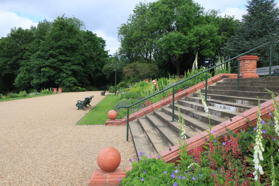 A landscaped garden area featuring a wide gravel pathway bordered by a well-maintained lawn with a few decorative orange spheres and black metal benches. To the right, a set of stone steps bordered by